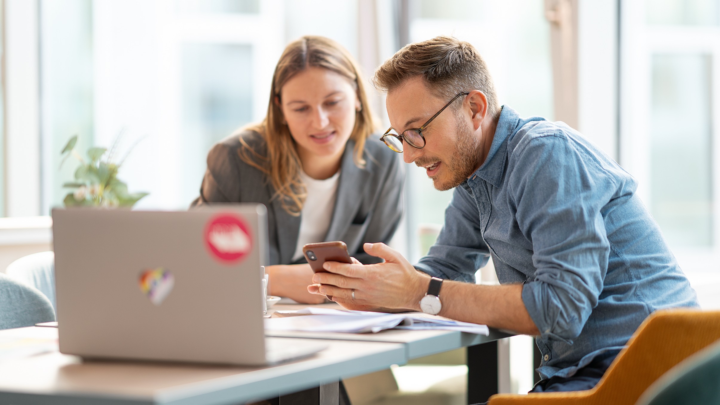 Zwei Personen sitzen am Schreibtisch, auf dem Dokumente liegen und ein Notebook steht und schauen auf ein Smartphone, das eine Person der anderen zeigt.