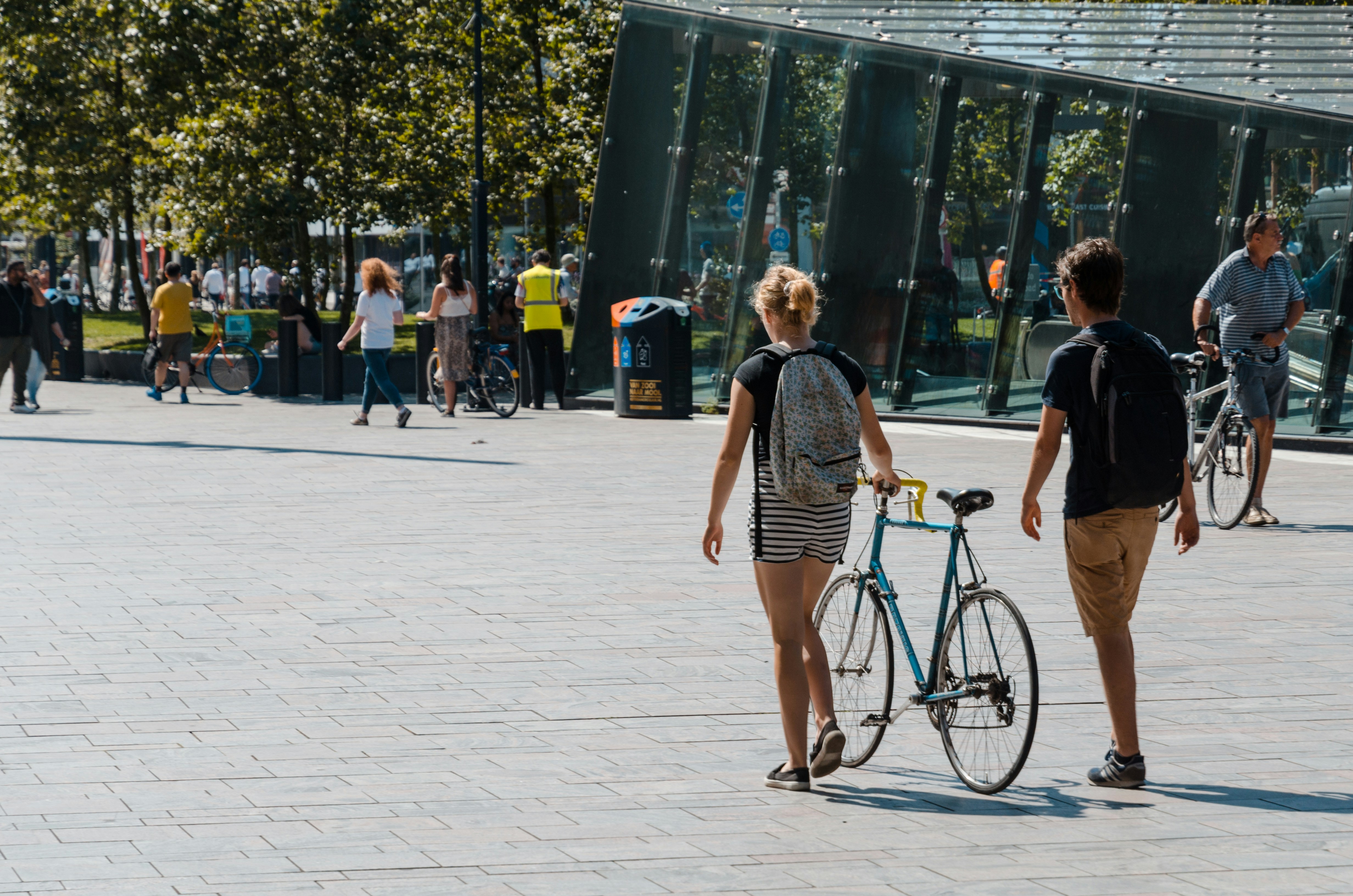 Zwei Personen mit Rucksäcken gehen von hinten fotografiert über einen großen, gepflasterten Platz, während eine von ihnen ein blaues Fahrrad neben sich her schiebt.