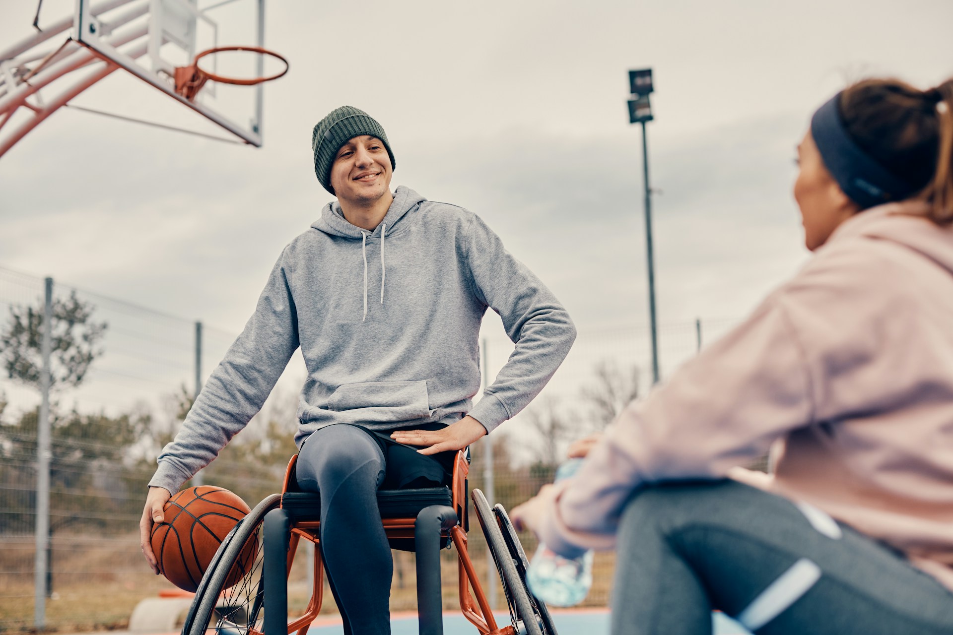 Rollstuhlfahrer unterhält sich mit Basketball unter dem Arm auf dem Sportplatz mit einer weiblichen Person. 