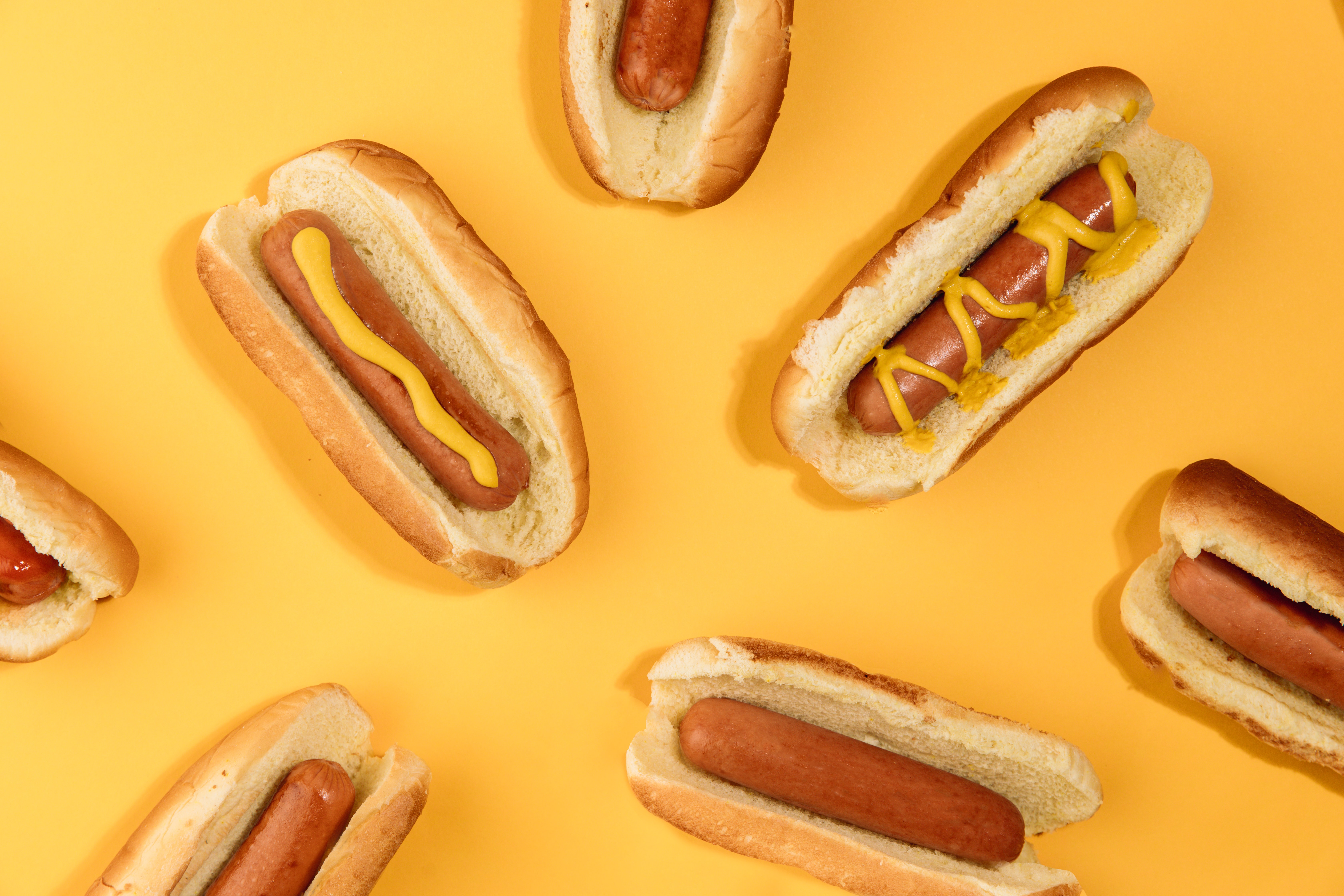Many hotdogs placed artfully over a yellow background. Two of the hotdogs have yellow mustard on them, in slightly different squiggle patterns.