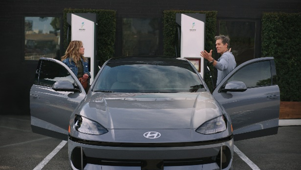 Sosie Bacon steht links, Kevin Bacon rechts jeweils in der geöffneten Vordertür eines Hyundai Ioniq 6. Sie sind einander zugewandt, er hat beide Arme ausgebreitet. Im Hintergrund stehen Ladesäulen. Szene aus einem Werbespot