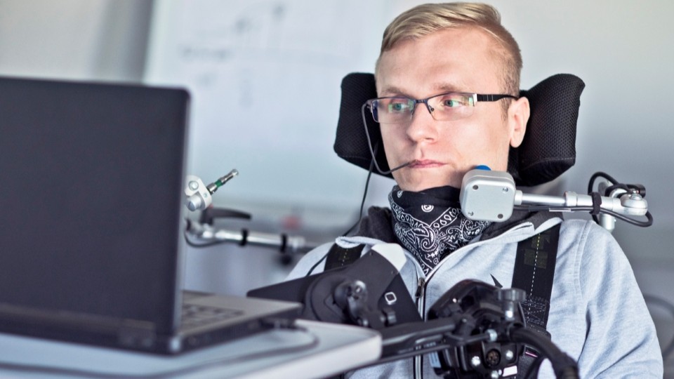 Mann mit körperlicher Einschränkung arbeitet mithilfe einer Spezialsteuerung am Laptop in einem barrierefreien Büro.