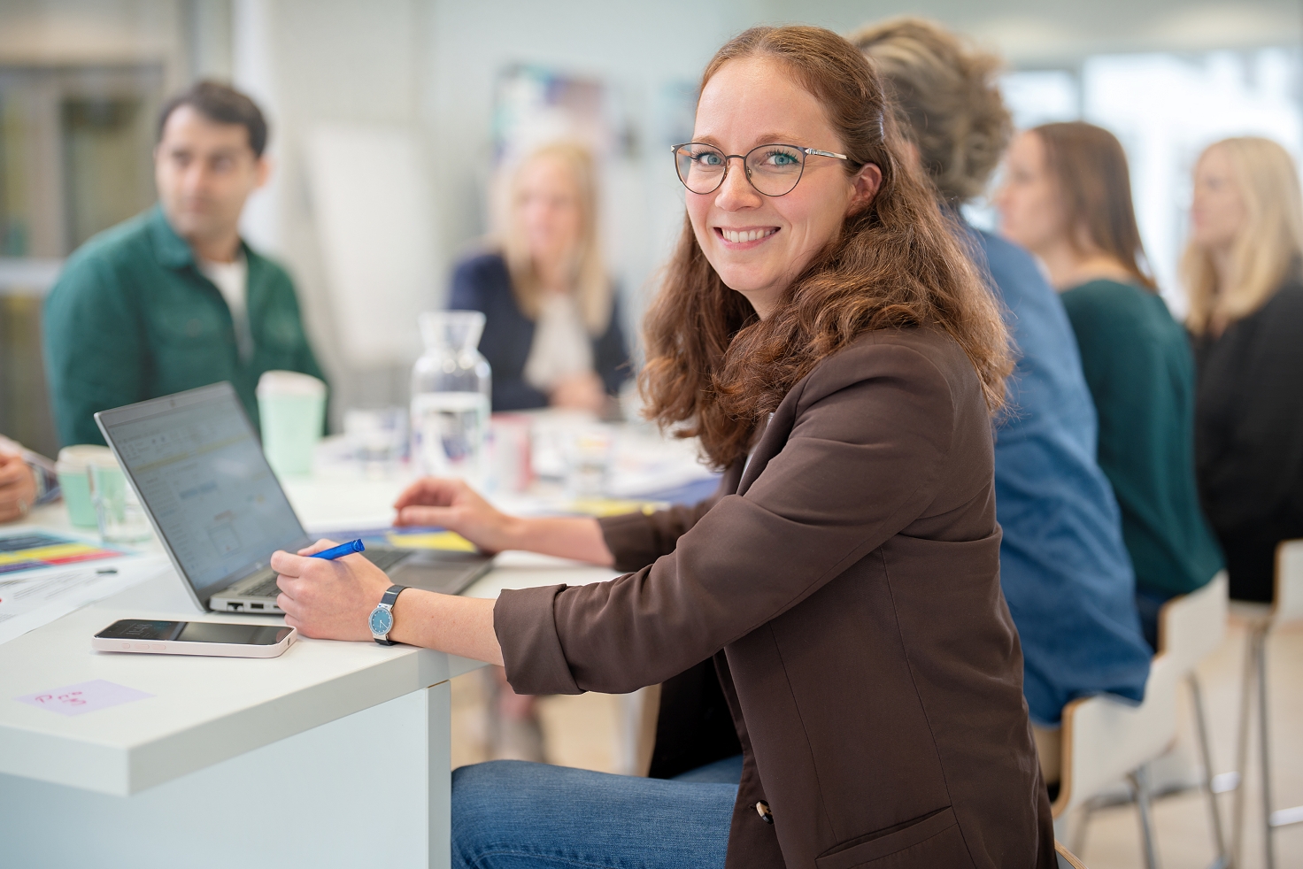 Frau sitzt an einem Besprechungstisch in einem modernen Büro und schaut lächeln in die Kamera, im Hintergrund sitzen mehrere weitere Personen und unterhalten sich