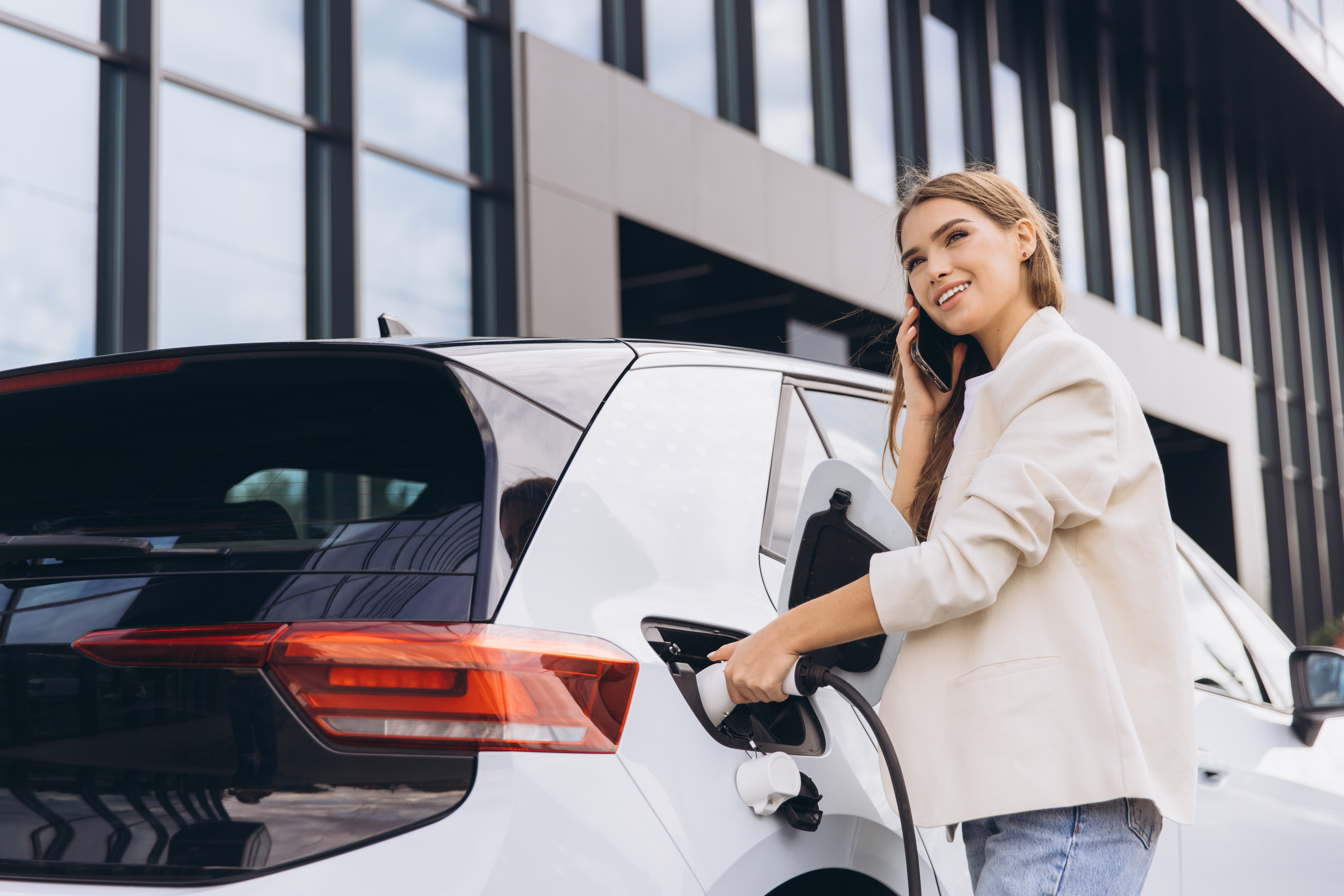Eine lächelnde Geschäftsfrau lädt ihr Elektroauto an einer modernen Ladestation auf, während sie telefoniert,