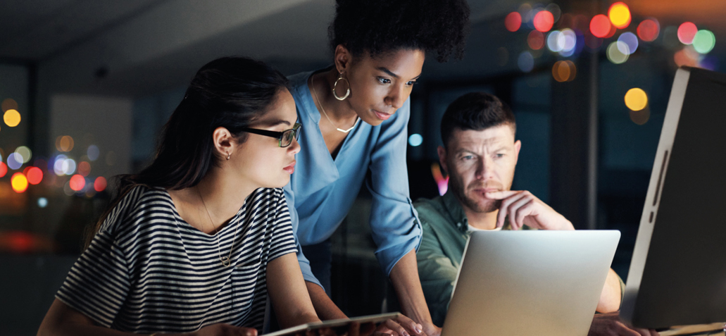 Diverse group of young executives crowd around a computer screen, collaborating into the late evening