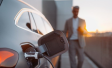 Die rechte Seite eines Elektroautos von hinten betrachtet, in dessen Ladevorrichtung ein Ladekabel steckt. Im Hintergrund ist ein Mensch vor einem Sonnenuntergang unscharf zu erkennen.