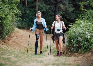 Zwei lächelnde Frauen wandern mit Trekkingstöcken einen von Bäumen gesäumten Pfad entlang, wobei eine der beiden eine schwarze Beinprothese trägt.