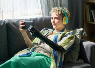 Eine Person mit lockigem Haar und Kopfhörern liegt entspannt auf einem Sofa in einem hellen Wohnzimmer und betrachtet ein Smartphone, das sie mit ihrer bionischen Armprothese hält.
