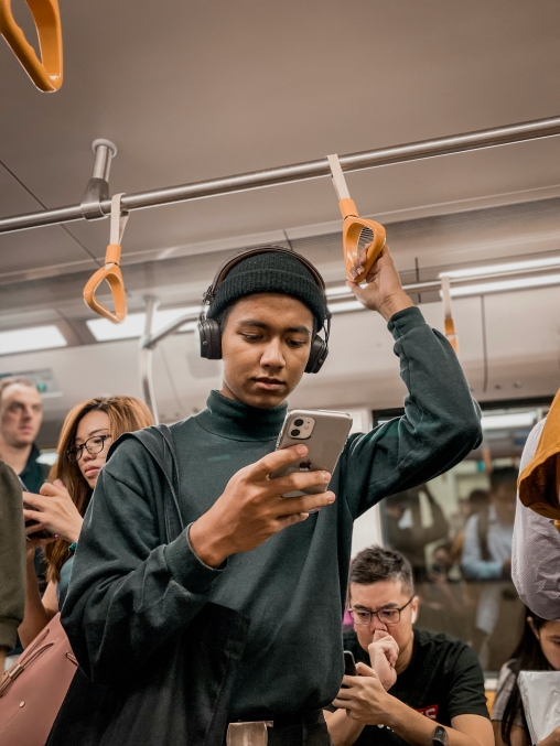 Eine Person mit Mütze und Kopfhörern steht in einer U-Bahn, hält sich an einer Schlaufe fest und blickt auf ein Smartphone, während im Hintergrund weitere Fahrgäste zu sehen sind.