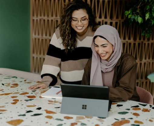 two women, one stood and one sitting, looking at a laptop