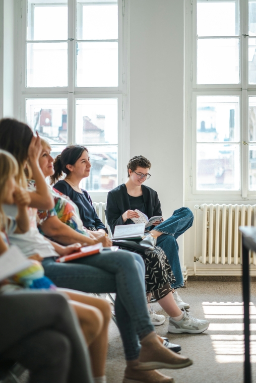 Eine Gruppe von Personen sitzt in einer Reihe in einem hellen, lichtdurchfluteten Raum. Die Atmosphäre erinnert an einen Workshop, ein Seminar oder eine Unterrichtsstunde.