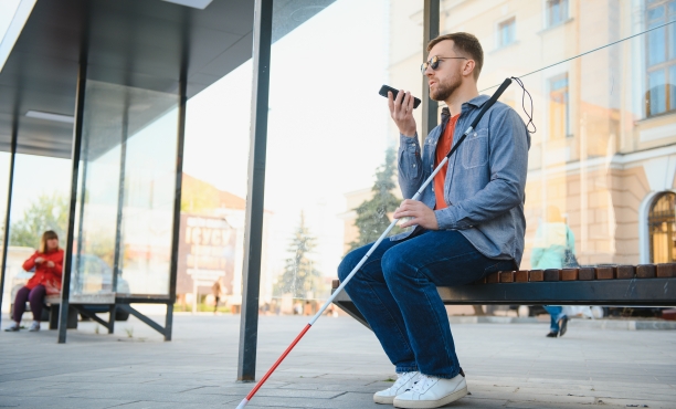 Ein sehbehinderter Mann mit Sonnenbrille und Blindenstock sitzt auf einer Bank an einer Bushaltestelle und spricht in sein Smartphone.