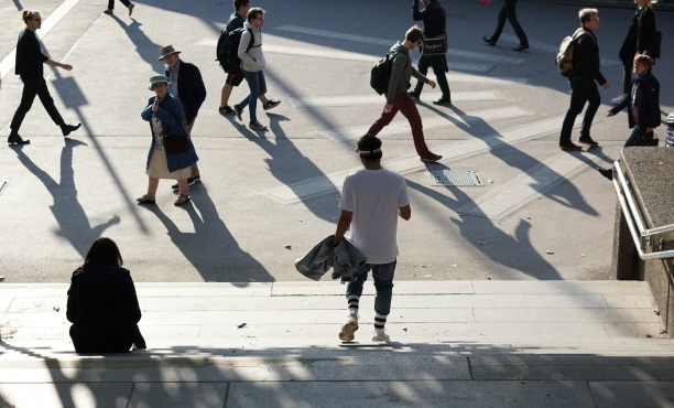Mehrere Passanten überqueren einen sonnenbeschienenen städtischen Platz und werfen lange, markante Schatten auf den Boden, während eine Person im Vordergrund eine breite Treppe hinabgeht.