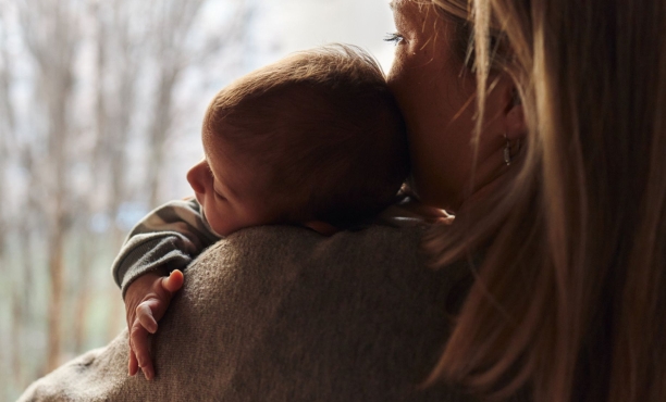 Rear view of a woman holding a sleeping baby on her shoulder near a window, with soft natural light creating a warm and comforting scene.