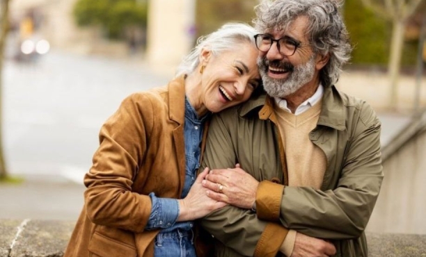 An older couple smile and laugh together outdoors, with the woman leaning her head on the man’s shoulder as they hold hands.