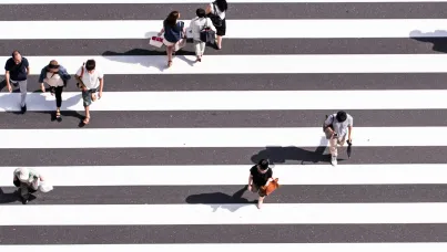 Draufsicht auf einen Zebrastreifen, über den mehrere Menschen in verschiedene Richtungen gehen, begleitet von ihren Schatten auf dem Straßenbelag