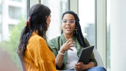 two women sat having a conversation, one holds a tablet
