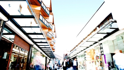 Shoppers walking through a modern outdoor shopping arcade with glass storefronts.
