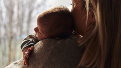Rear view of a woman holding a sleeping baby on her shoulder near a window, with soft natural light creating a warm and comforting scene.