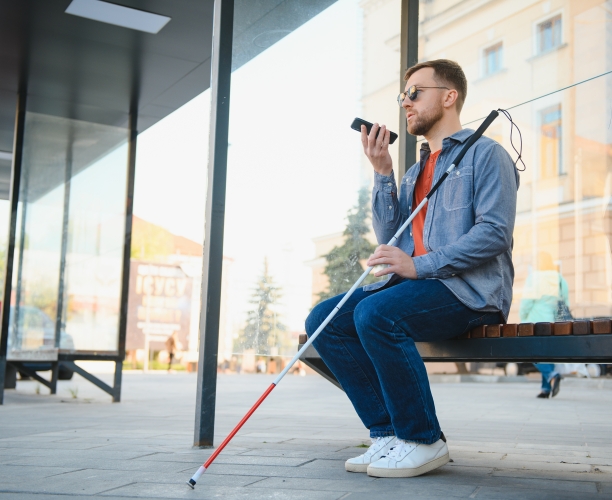Ein sehbehinderter Mann mit Sonnenbrille und Blindenstock sitzt auf einer Bank an einer Bushaltestelle und spricht in sein Smartphone.
