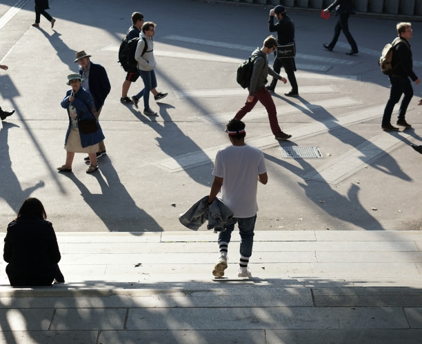 Mehrere Passanten überqueren einen sonnenbeschienenen städtischen Platz und werfen lange, markante Schatten auf den Boden, während eine Person im Vordergrund eine breite Treppe hinabgeht.