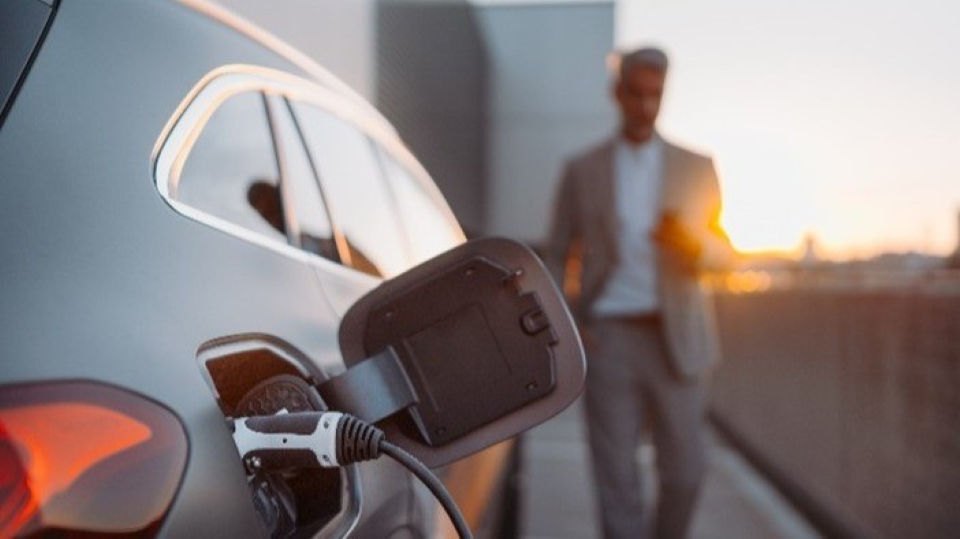 Die rechte Seite eines Elektroautos von hinten betrachtet, in dessen Ladevorrichtung ein Ladekabel steckt. Im Hintergrund ist ein Mensch vor einem Sonnenuntergang unscharf zu erkennen.