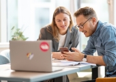 Clarissa Quiring und Peter Paul Plambeck, Marketing & Communications, Ipsos in Deutschland, sitzen vor einem aufgeklappten Laptop am Tisch und schauen mit interessierten Gesichtsausdrücken auf ein Smartphone, das Peter Paul Plambeck in seiner linken Hand hält.